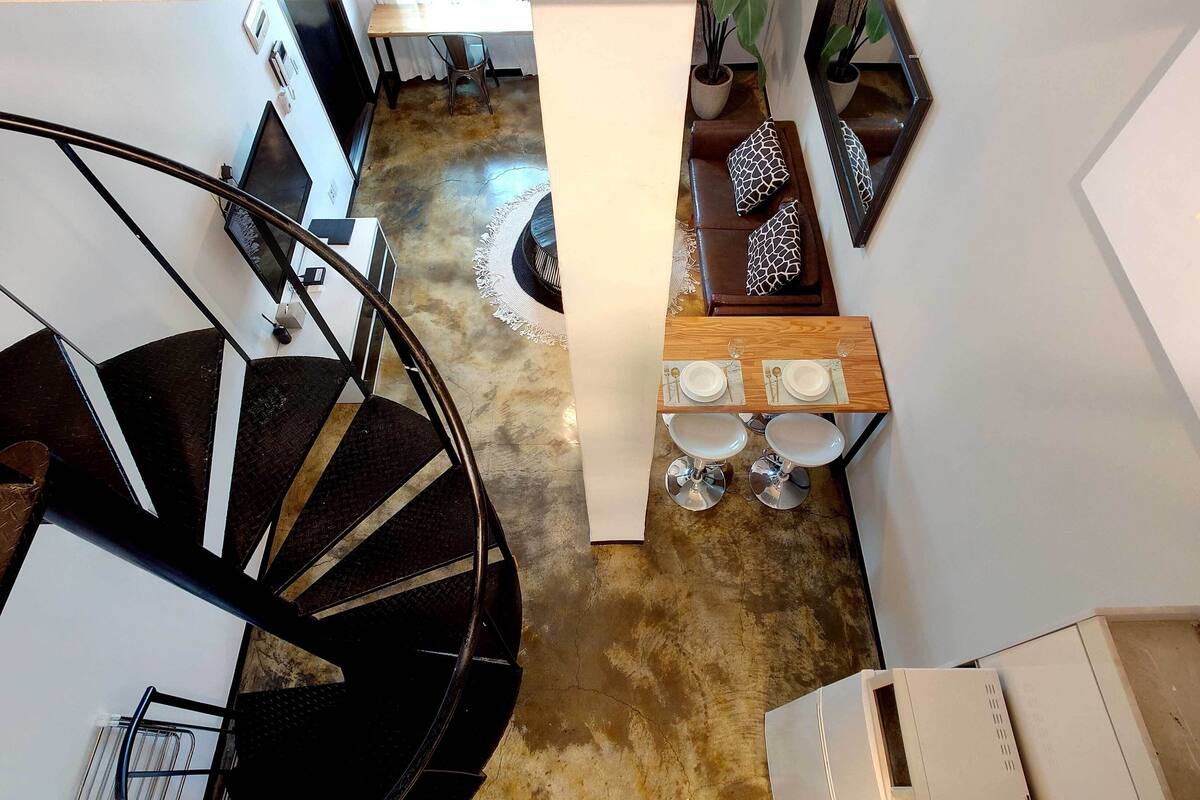 Un interior moderno con una escalera espiral, un comedor con una mesa y sillas de madera, y un sofá con cojines estampados.