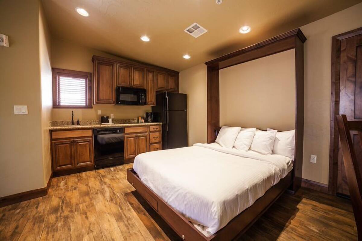 A bedroom with a large bed, wooden flooring, a kitchen with wooden cabinets, a refrigerator, and a microwave.
