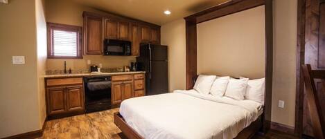 A bedroom with a large bed, wooden flooring, a kitchen with wooden cabinets, a refrigerator, and a microwave.