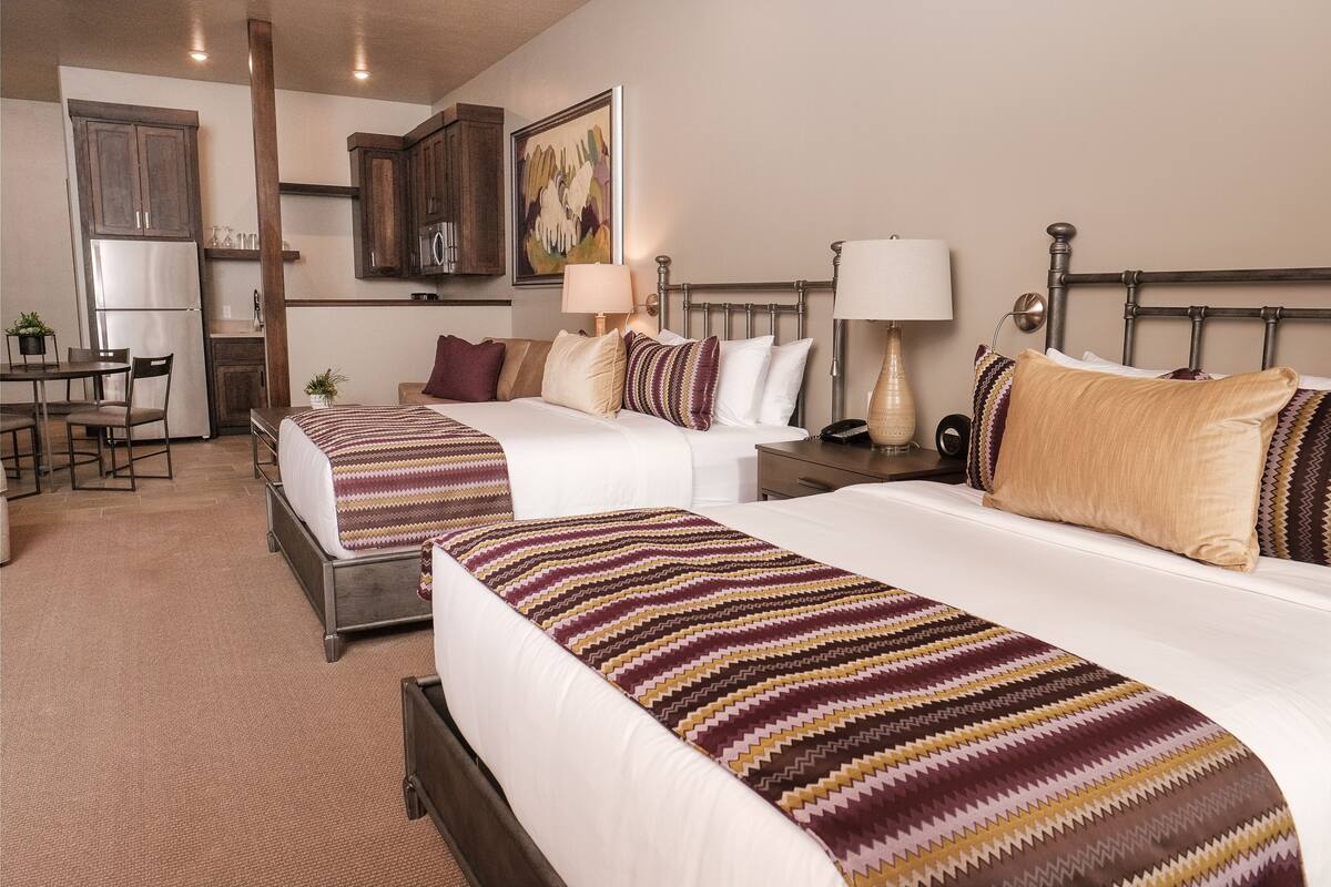 A hotel room with two beds, a kitchenette, a dining area, and a painting on the wall.