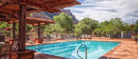 A pool area with a wooden pergola, lounge chairs, and a view of mountains.