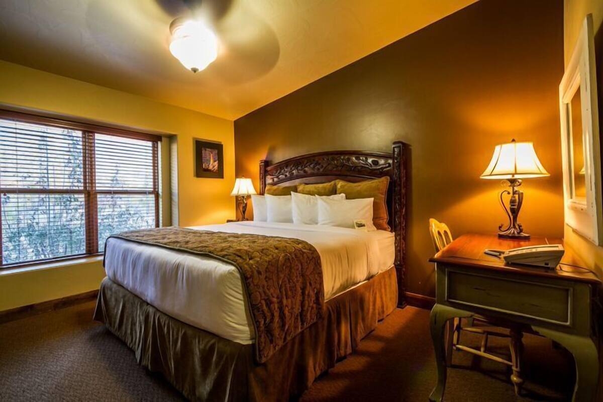 A hotel room with a large bed, a desk with a lamp, and a window with blinds.