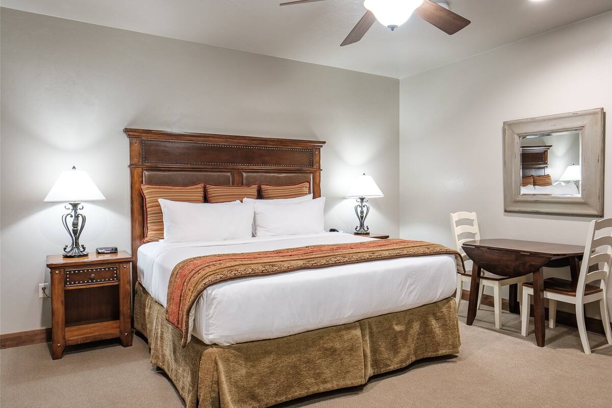 A bedroom with a large bed, two bedside tables, a wooden headboard, a table with chairs, and a ceiling fan.