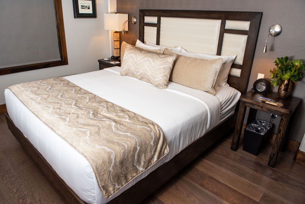 A neatly made bed with a marble-patterned bedspread and pillows, a wooden headboard, a bedside lamp, a small table with a plant, and a trash bin.