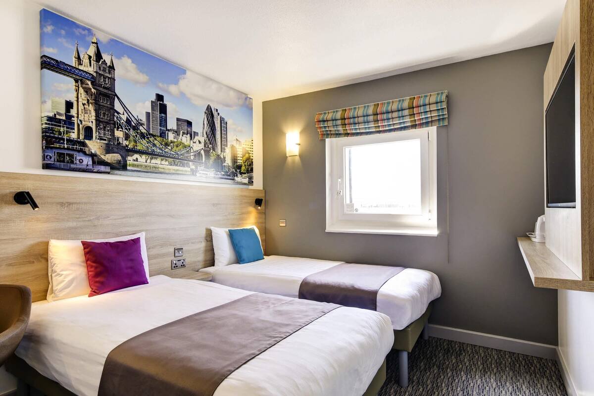 Une chambre d’hôtel avec deux lits, un tableau représentant un paysage urbain, une fenêtre agrémentée d’une lambrequin à carreaux et un téléviseur fixé au mur.