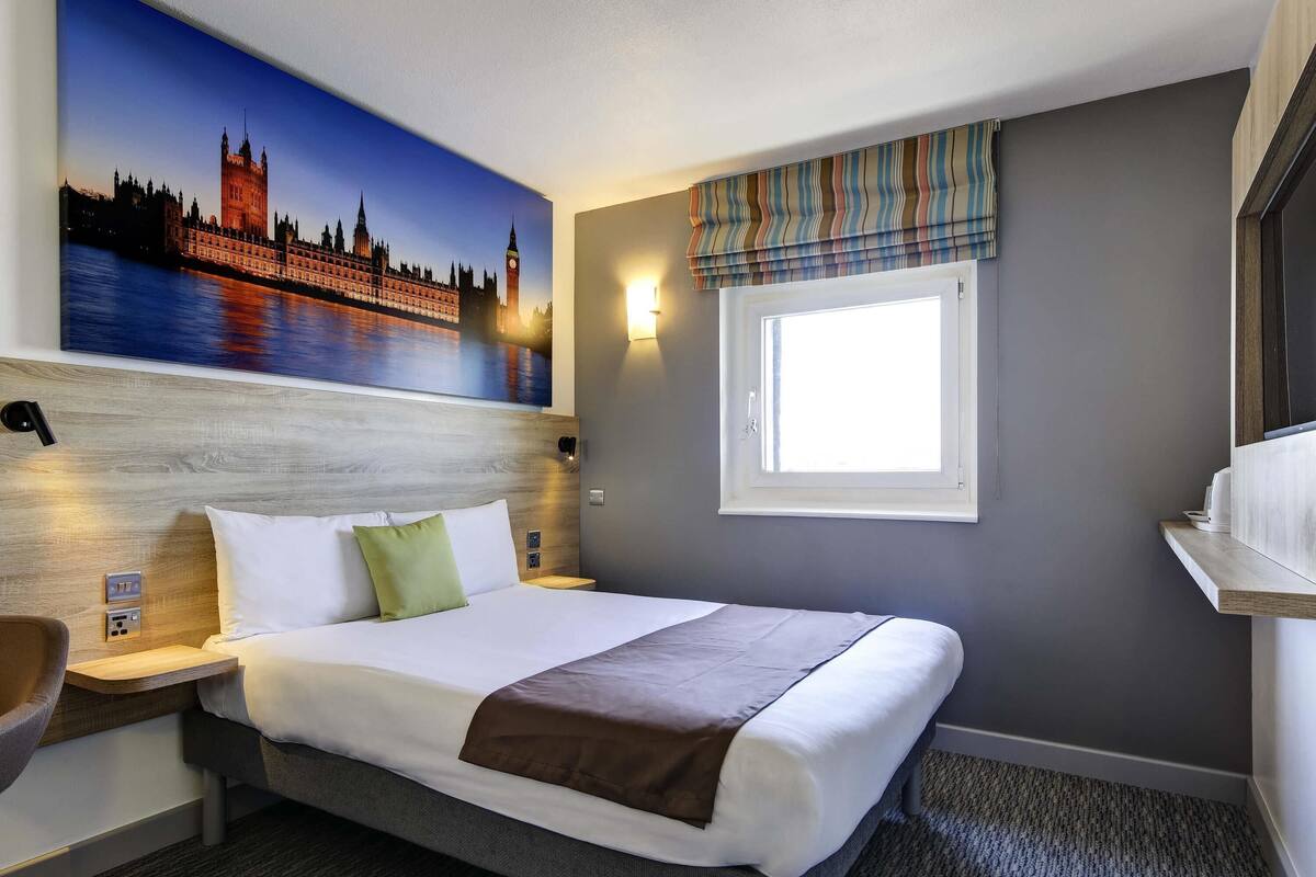 Une chambre d’hôtel dotée d’un grand lit, d’une tête de lit ornée d’une fresque représentant un paysage urbain, d’une table de chevet avec une lampe, d’une fenêtre offrant une vue et d’un petit coin salon.
