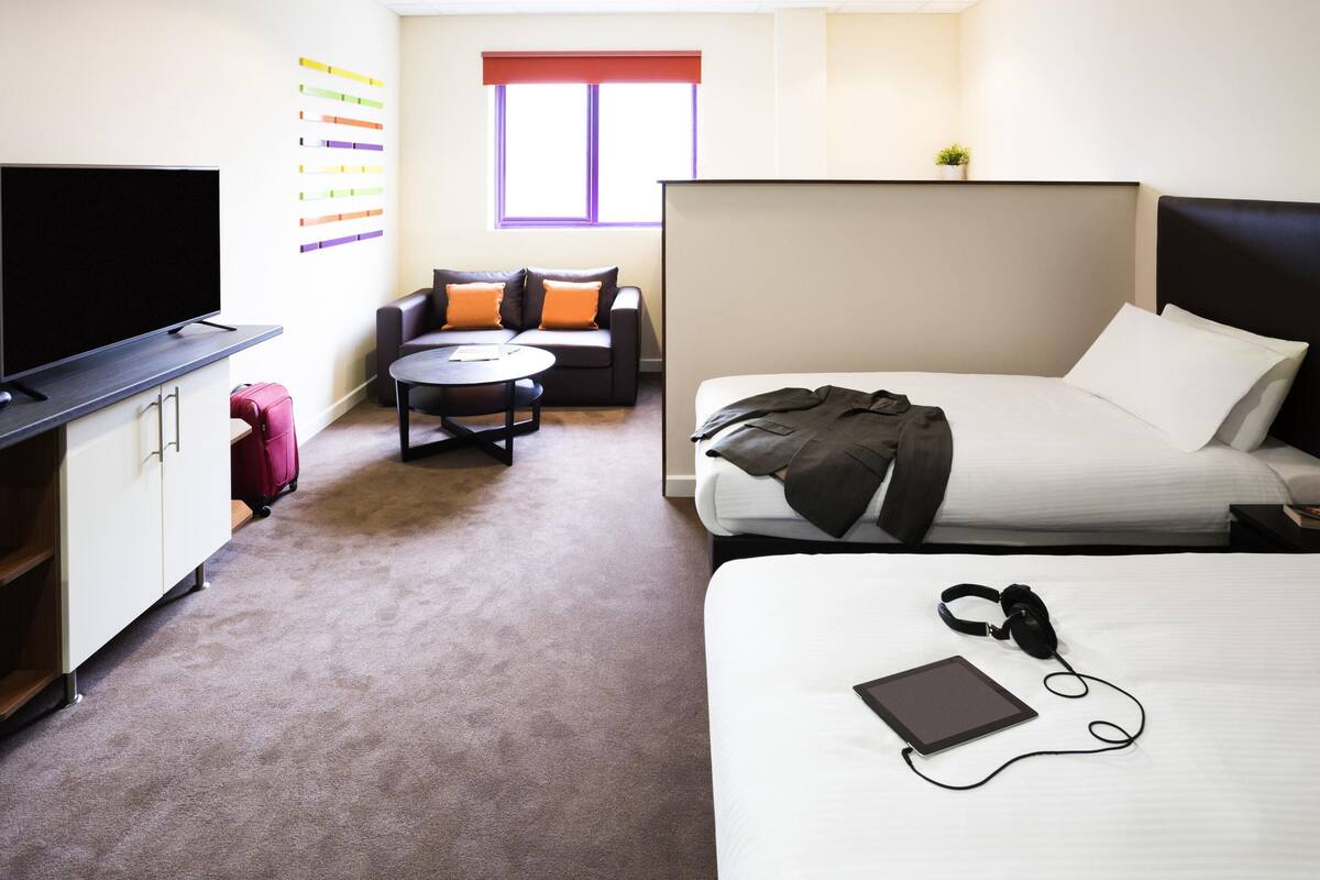 Une chambre d’hôtel avec un lit, un bureau avec une télévision, un canapé, une petite table ronde et une fenêtre.