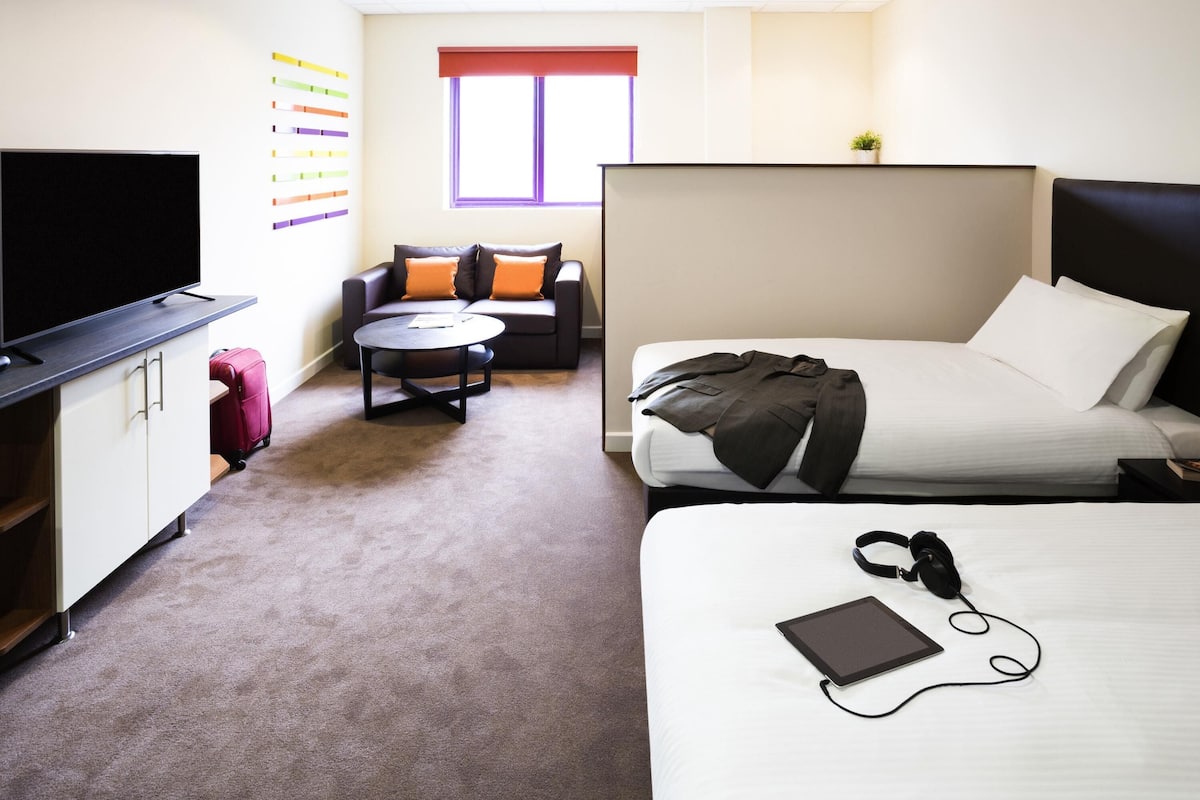 Une chambre dâhĂŽtel avec un lit, un bureau avec une tĂ©lĂ©vision, un canapĂ©, une petite table ronde et une fenĂȘtre.