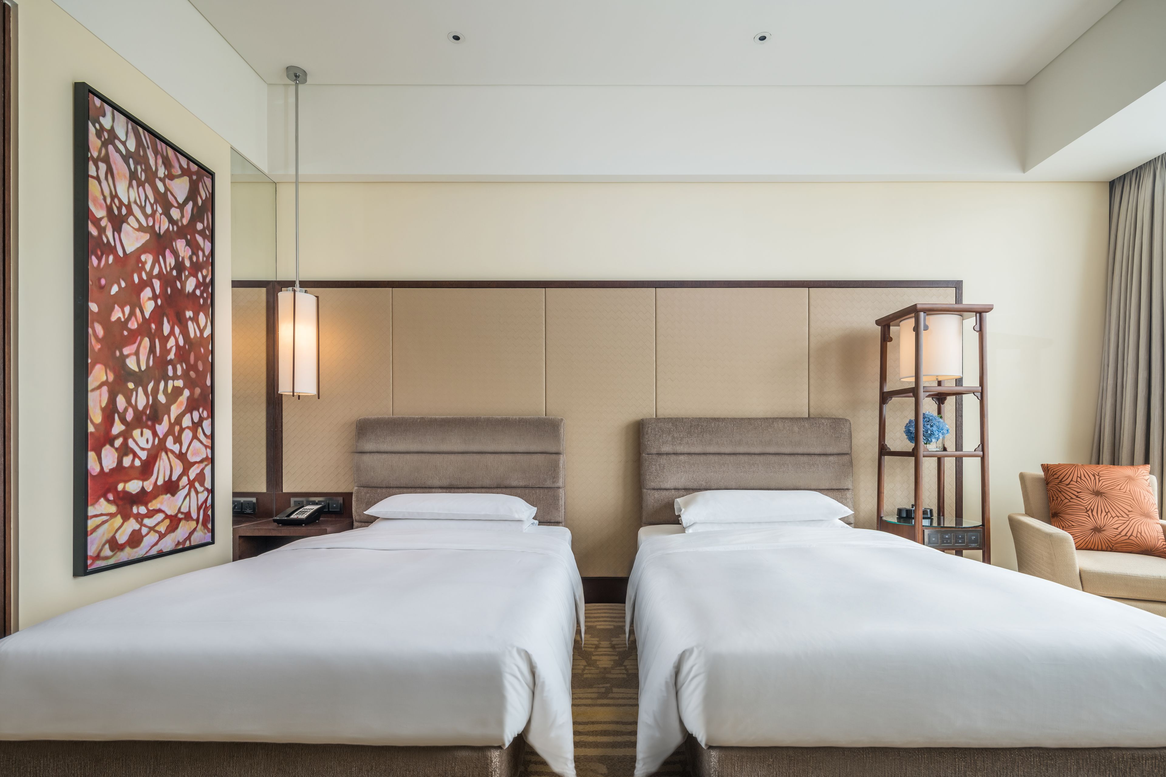 Habitación de hotel con dos camas, un cuadro abstracto de gran tamaño, una mesita de noche con lámpara, un mueble pequeño y una silla con cojín.