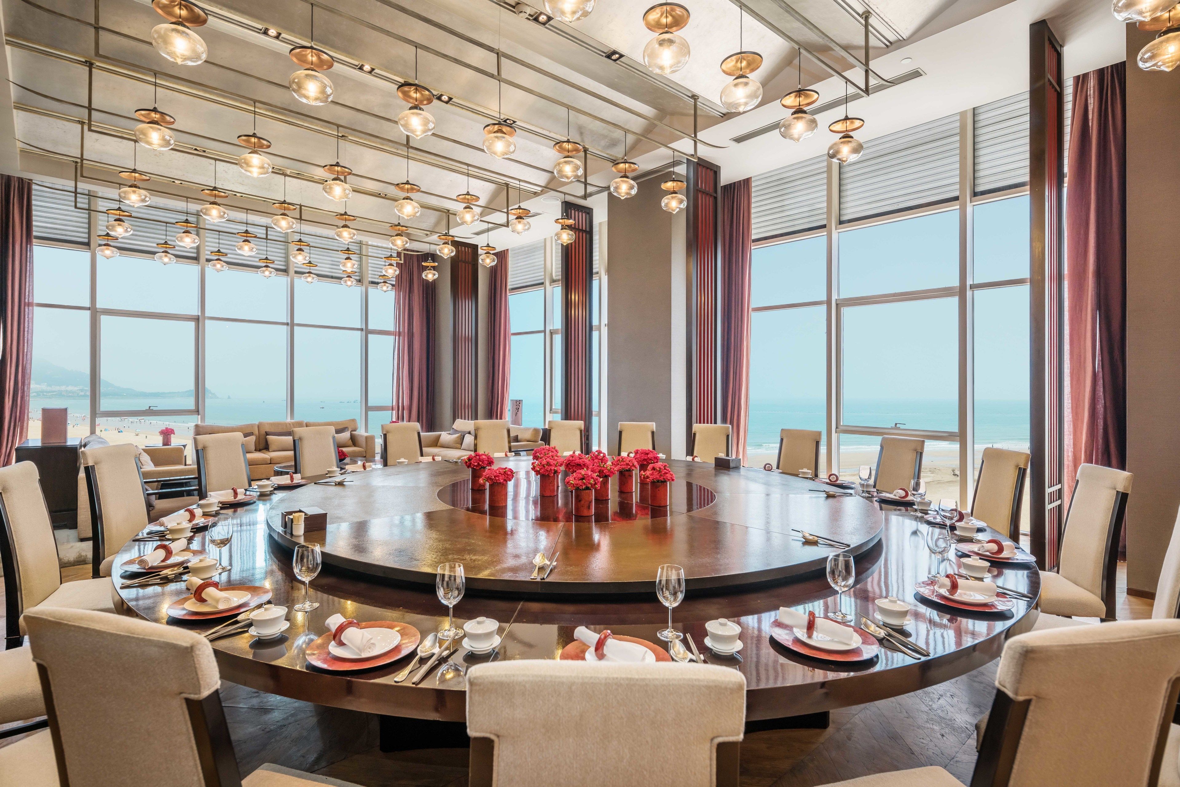 A round conference table with a wooden surface, surrounded by beige chairs, set for a meeting with floral centerpieces and a view of the sea through large windows.