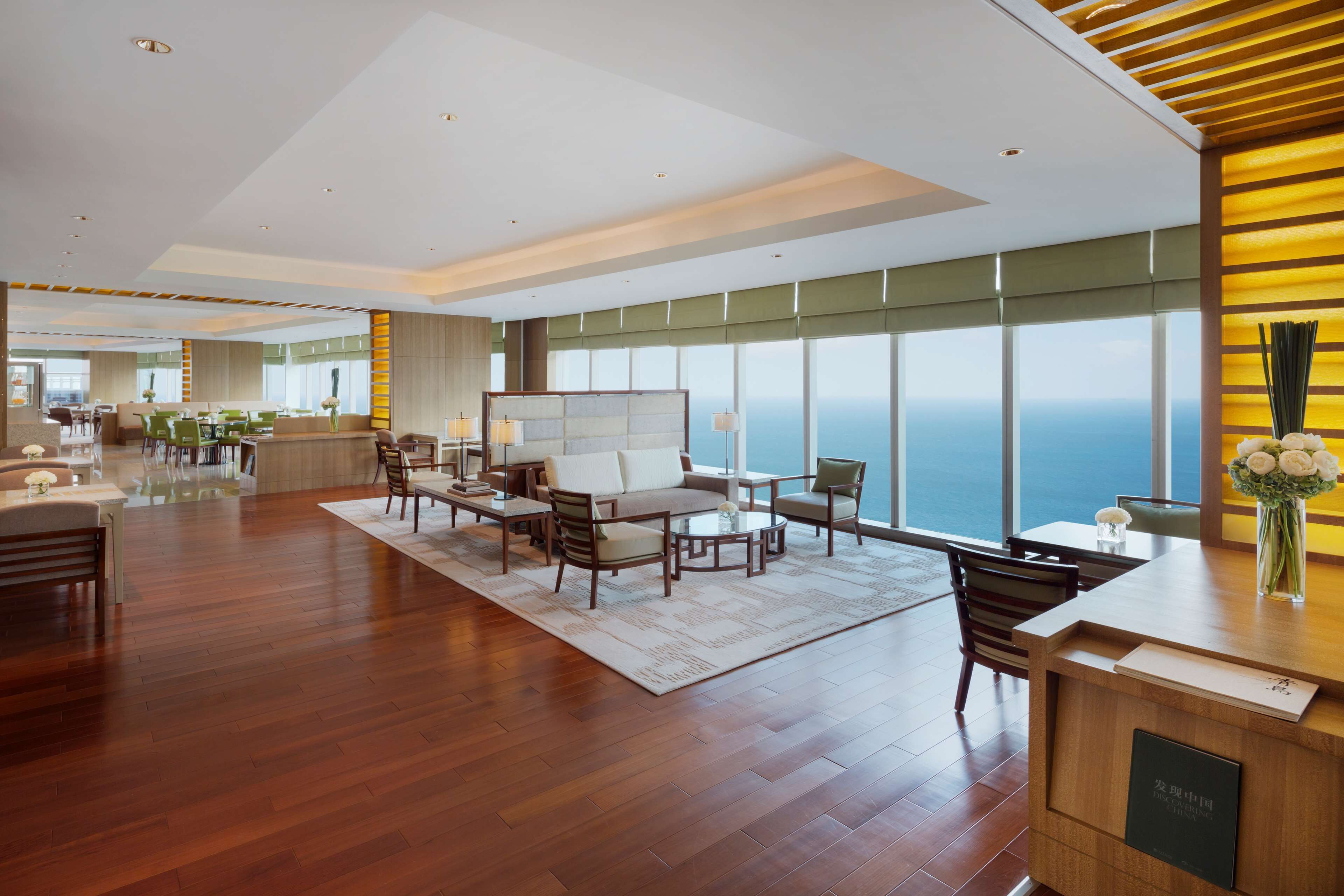 A spacious hotel lobby with a wooden reception desk, comfortable seating area, and large windows offering a view of the ocean.