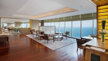 A spacious hotel lobby with a wooden reception desk, comfortable seating area, and large windows offering a view of the ocean.