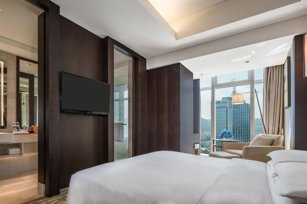 Ett modernt hotellrum med en stor säng, en tv som är monterad på väggen, ett badrum med handfat och spegel samt utsikt över stadsbilden genom stora fönster.