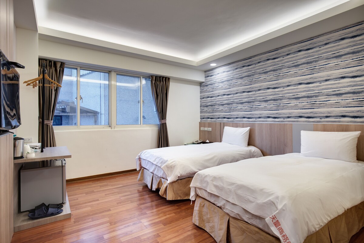A hotel room with two beds, a desk with a chair, a shoe rack, and a window with curtains.