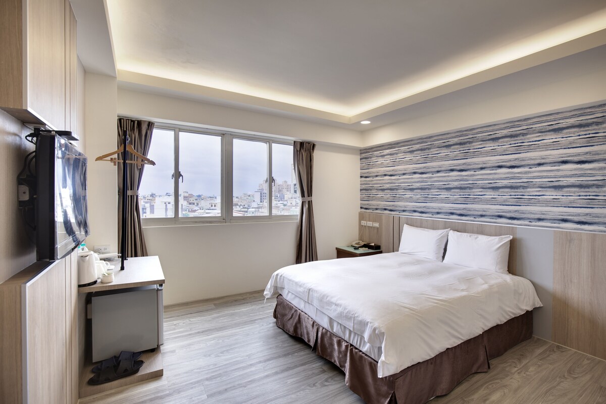 A modern hotel room with a large bed, a desk, and a view of the city through the window.