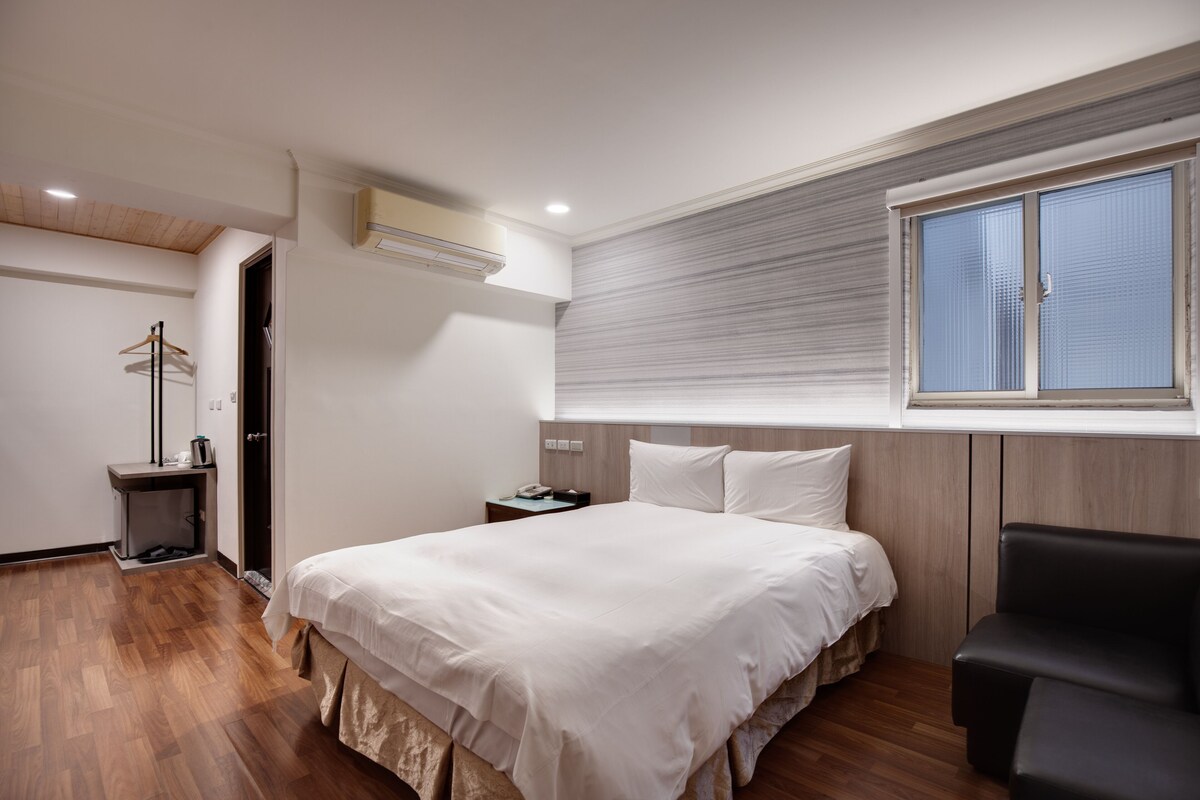 A modern hotel room with a large bed, a bedside table, a chair, and a window with blinds.