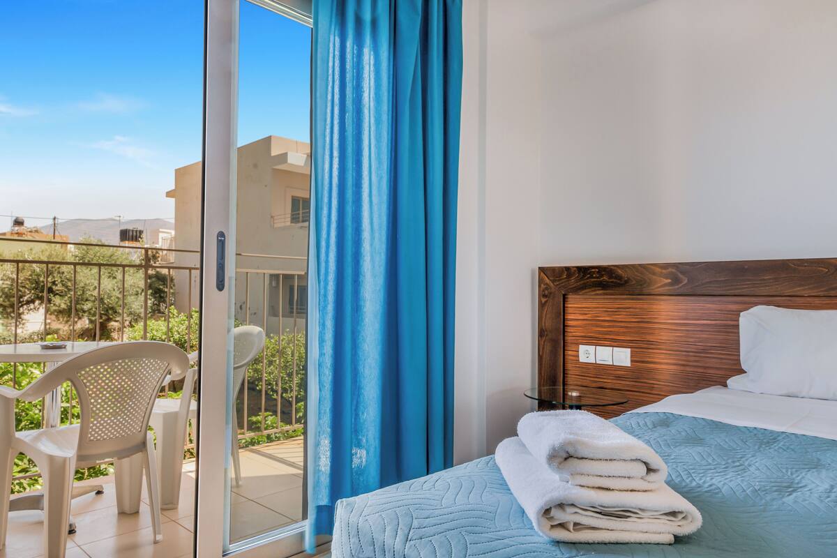 Una habitación de hotel con una cama, un balcón con cortinas azules, una silla blanca y una mesa con una silla.