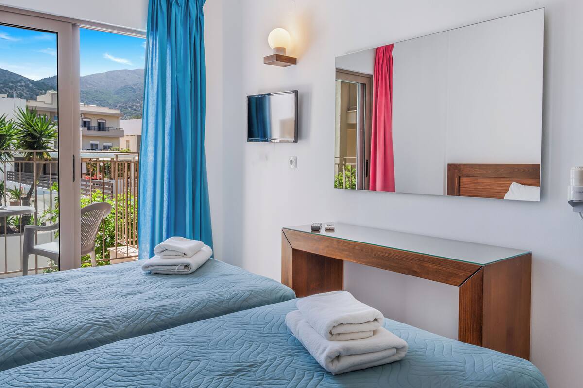 Habitación de hotel con dos camas, una mesita de noche de madera, un televisor y un balcón con vistas a las montañas.