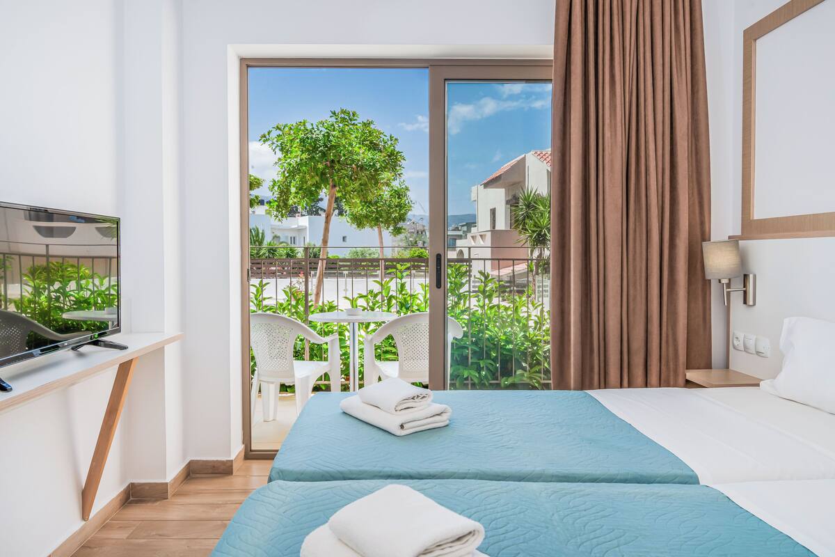 Habitación de hotel con cama, televisión, balcón con vistas a zonas verdes y edificios, y ventana con cortinas.