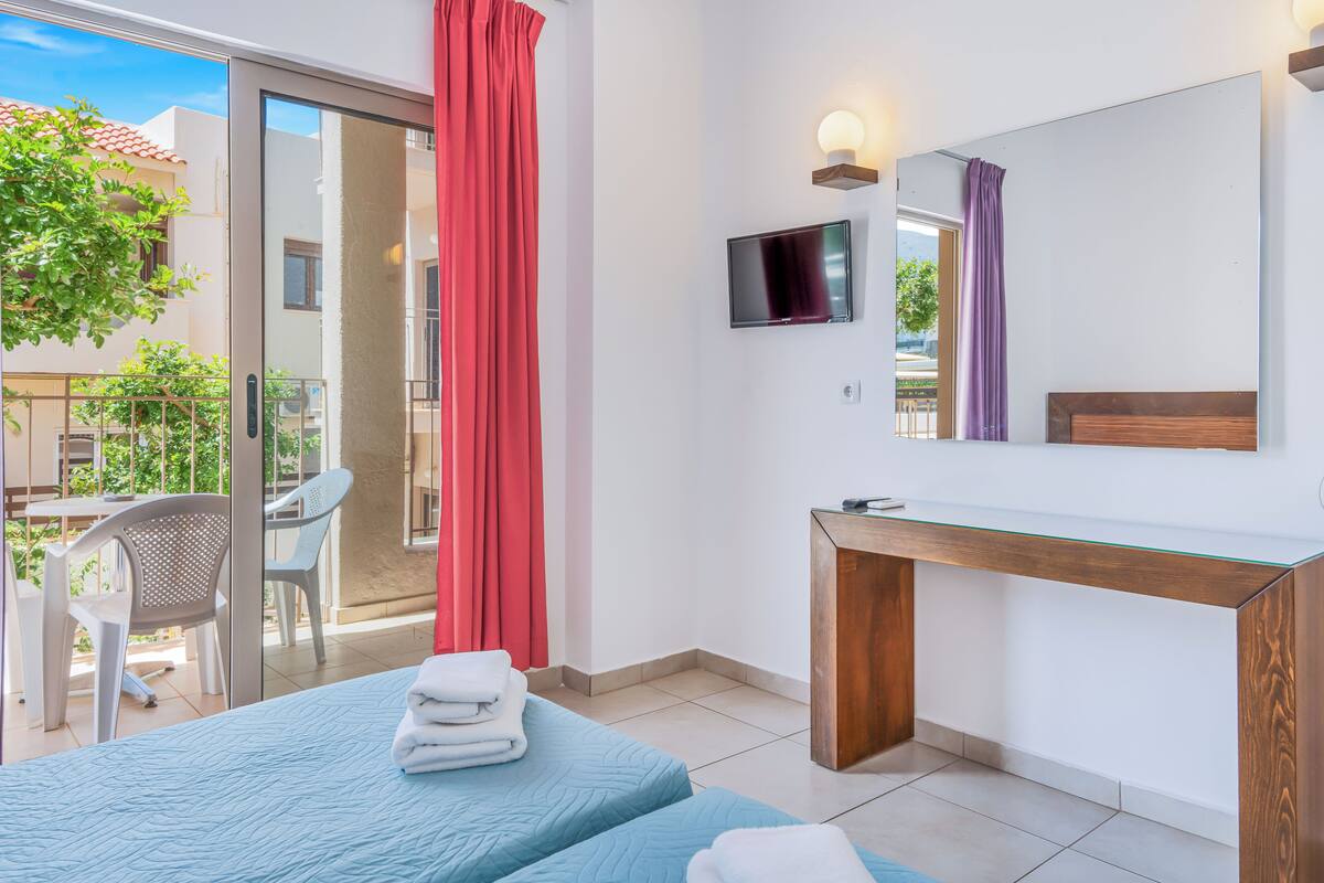 Habitación de hotel con cama, escritorio, televisor y balcón con sillas.