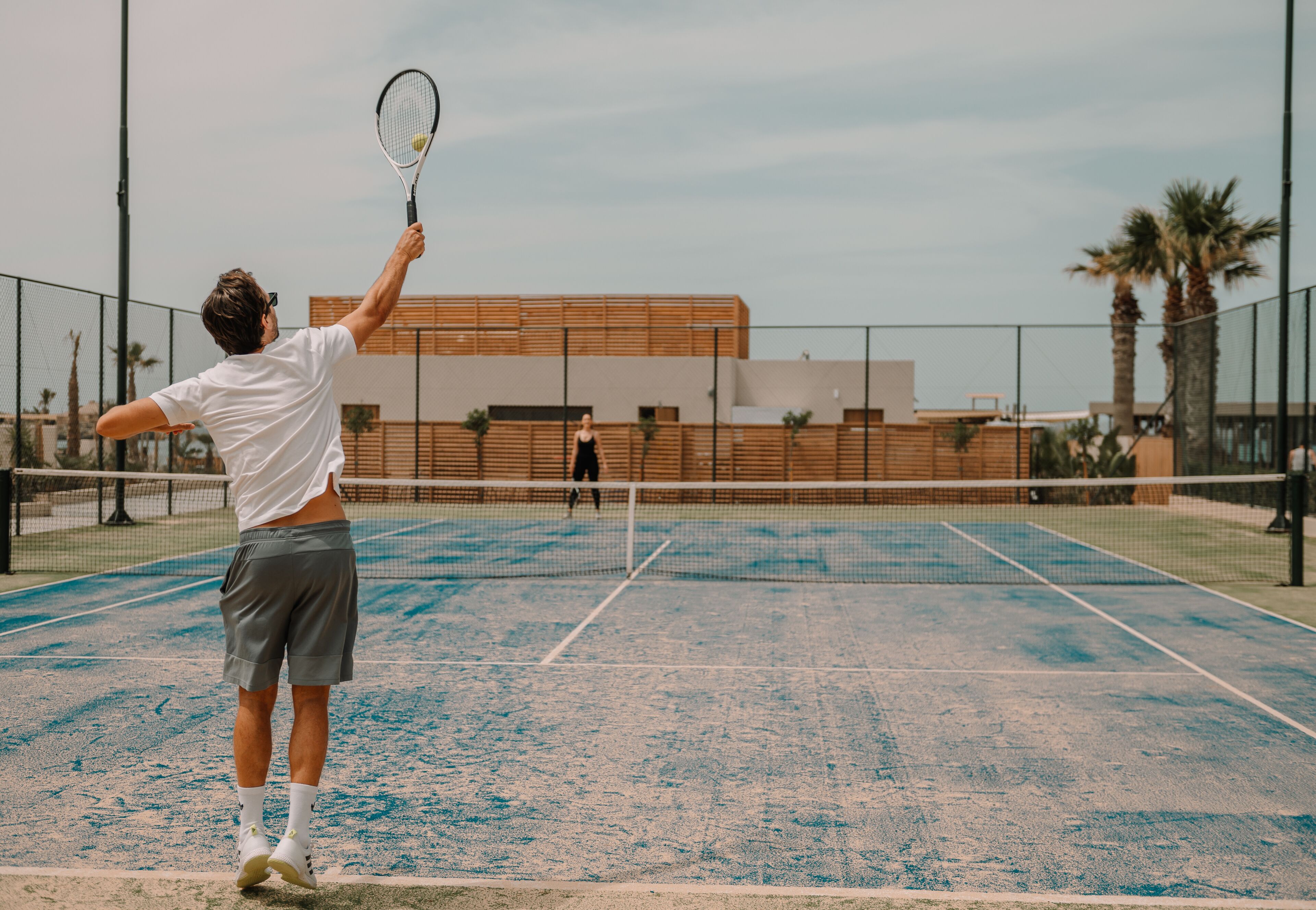 Una cancha de tenis con una red, un jugador con camisa blanca y shorts grises, y un edificio al fondo.