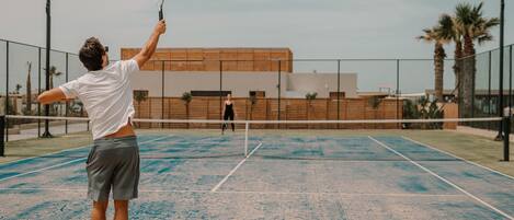 Una cancha de tenis con una red, un jugador con camisa blanca y shorts grises, y un edificio al fondo.