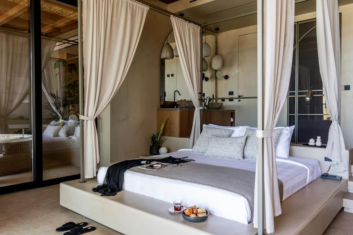 Ein modernes Schlafzimmer mit einem Himmelsbett, einem Nachttisch mit einem Glas Wasser und Snacks sowie Blick auf ein Badezimmer mit einer Badewanne und einem Waschbecken.