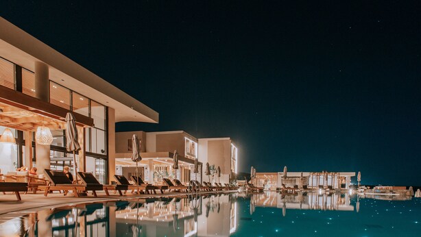 A nighttime view of a hotel pool area with sun loungers and umbrellas, reflecting the building's architecture. - Unique Blue! Resort and Villas Adults Only (Hersonissos)
