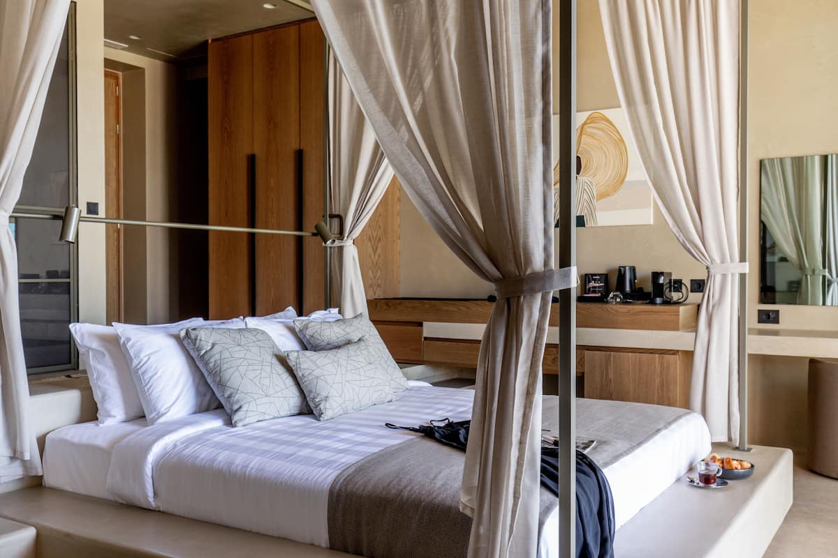 Ein modernes Hotelzimmer mit einem großen Bett, einem hölzernen Kopfteil, einem Nachttisch mit Lampe und Blick ins Zimmerinnere.