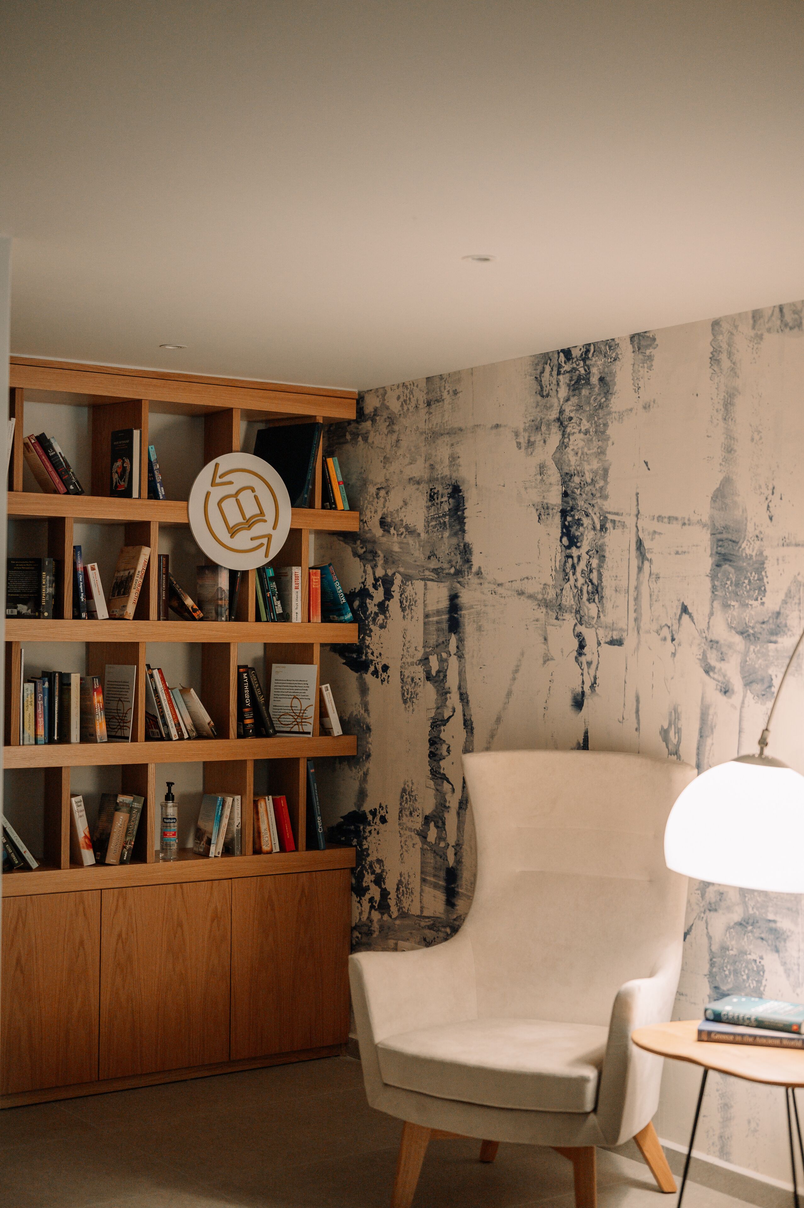 Una habitación con una estantería de madera llena de libros, un sillón blanco y una pequeña mesa con una lámpara.