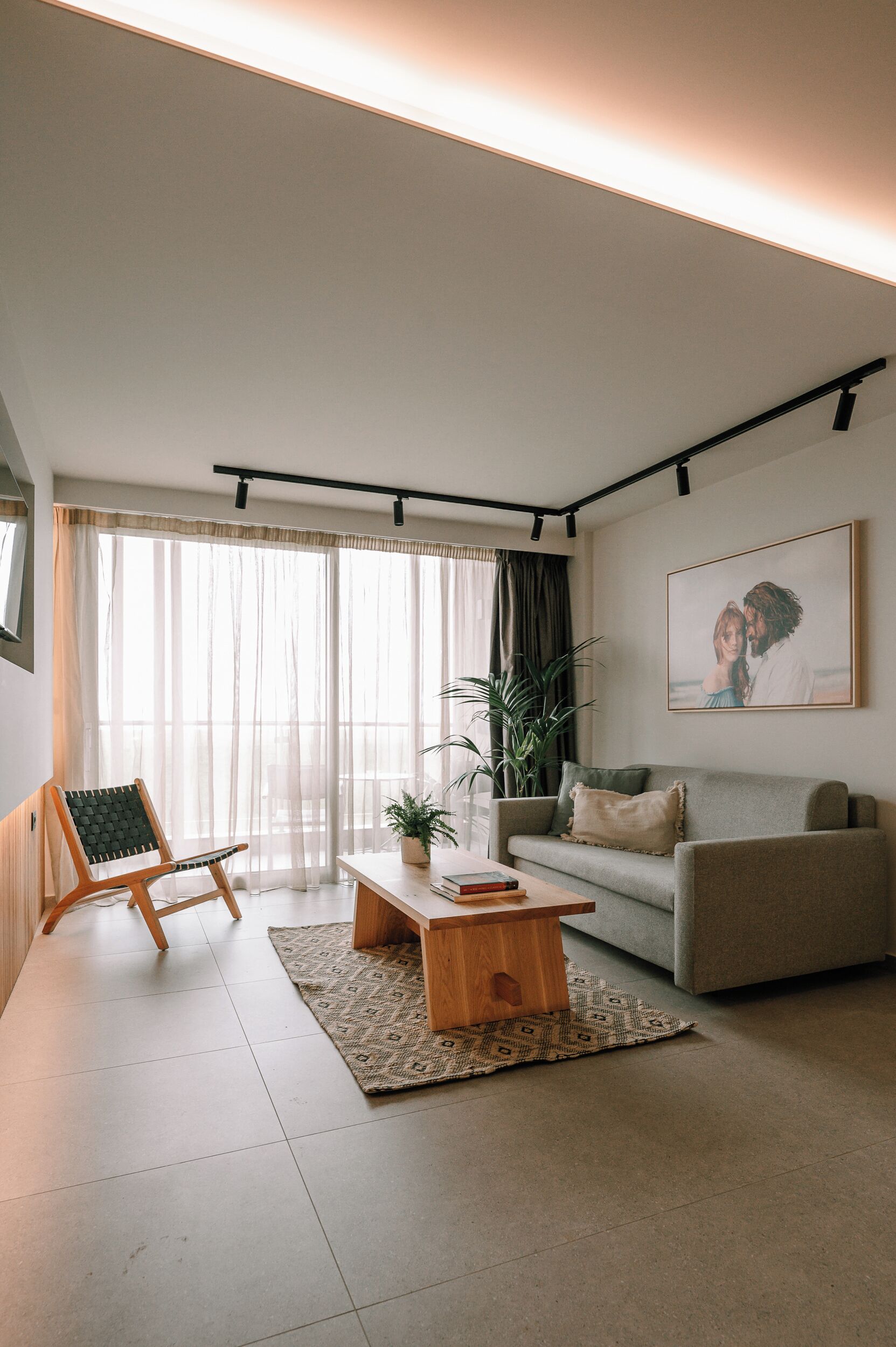 A modern living room with a sofa, a wooden coffee table, a chair, and a large window with sheer curtains.