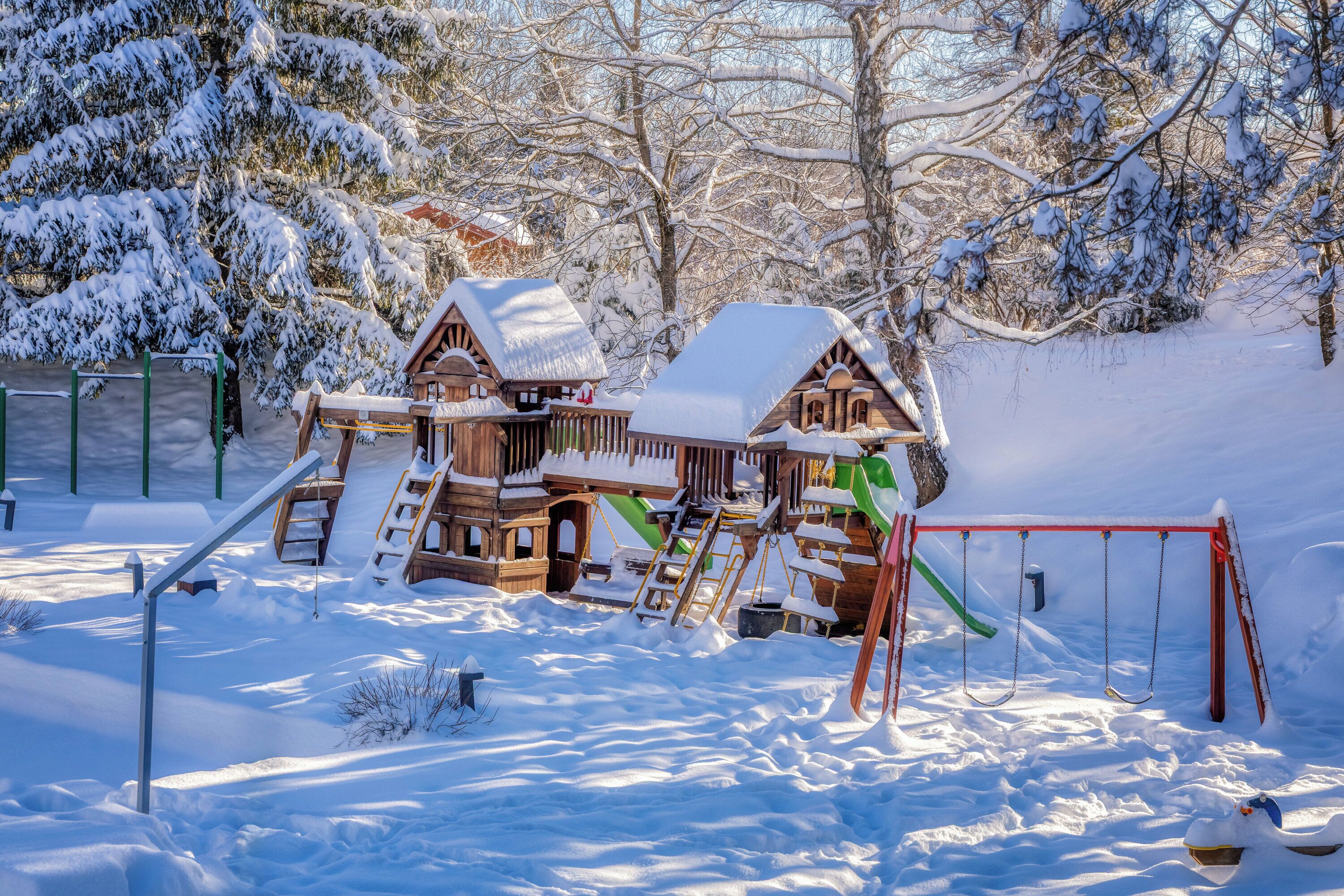 A wooden playhouse with a slide and swing set in a snowy outdoor area.