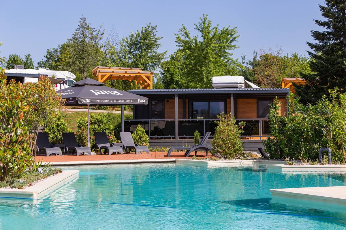 A modern building with a pool, lounge chairs, and a covered area with umbrellas.