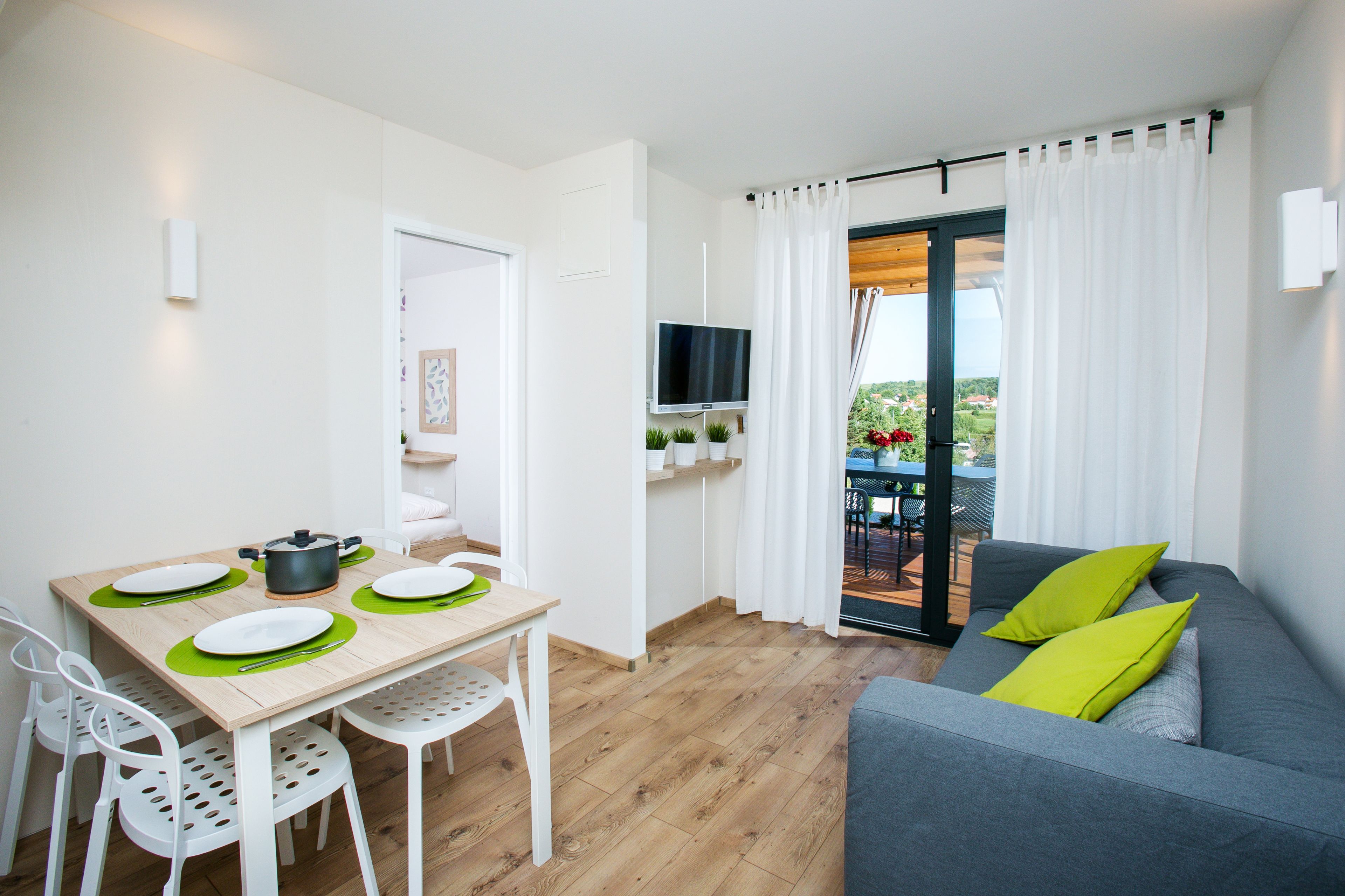 A modern living room with a grey sofa, a dining table with white chairs, and a balcony with a TV.