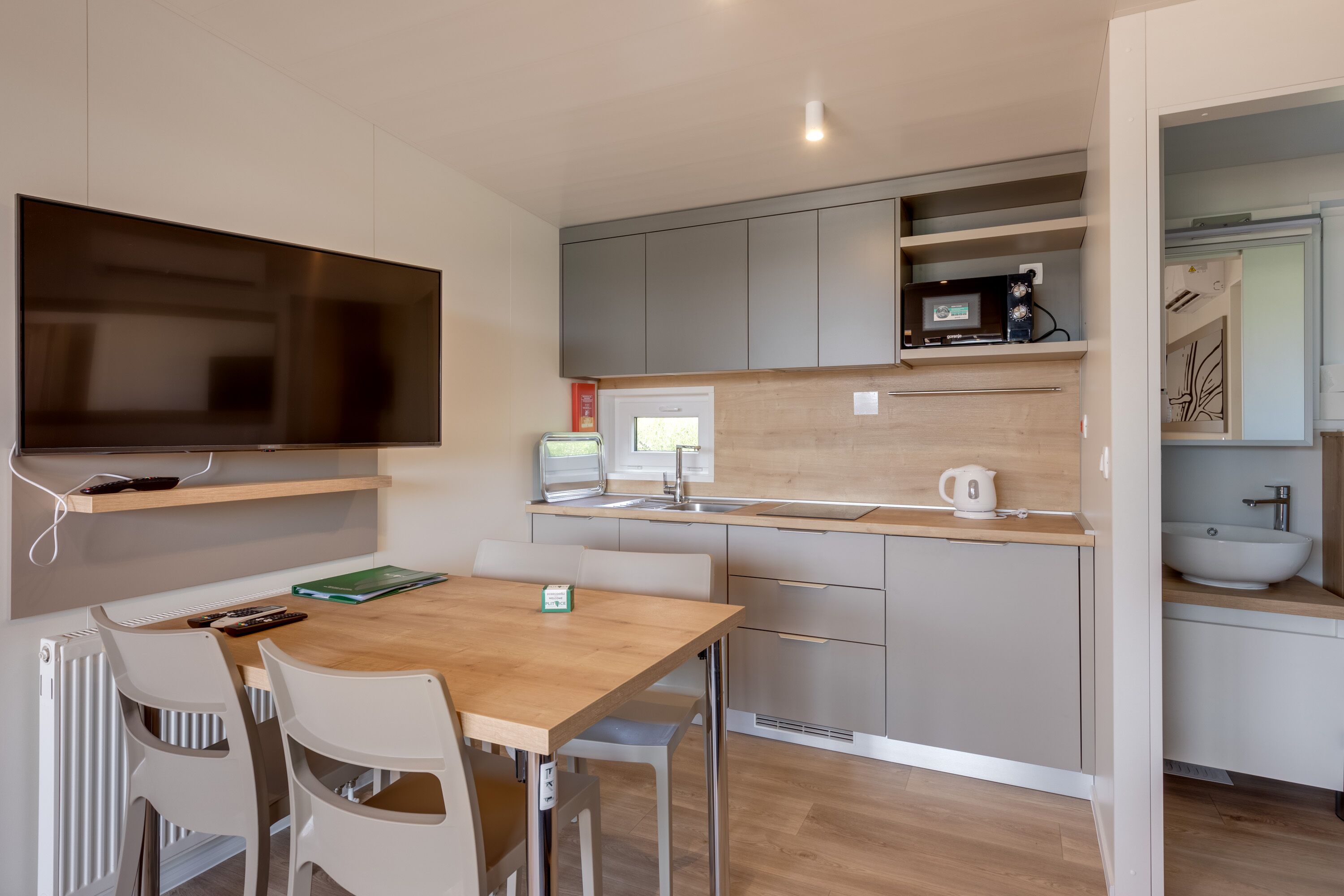 A modern kitchen with a wooden table, white chairs, a built-in TV, and grey cabinets.