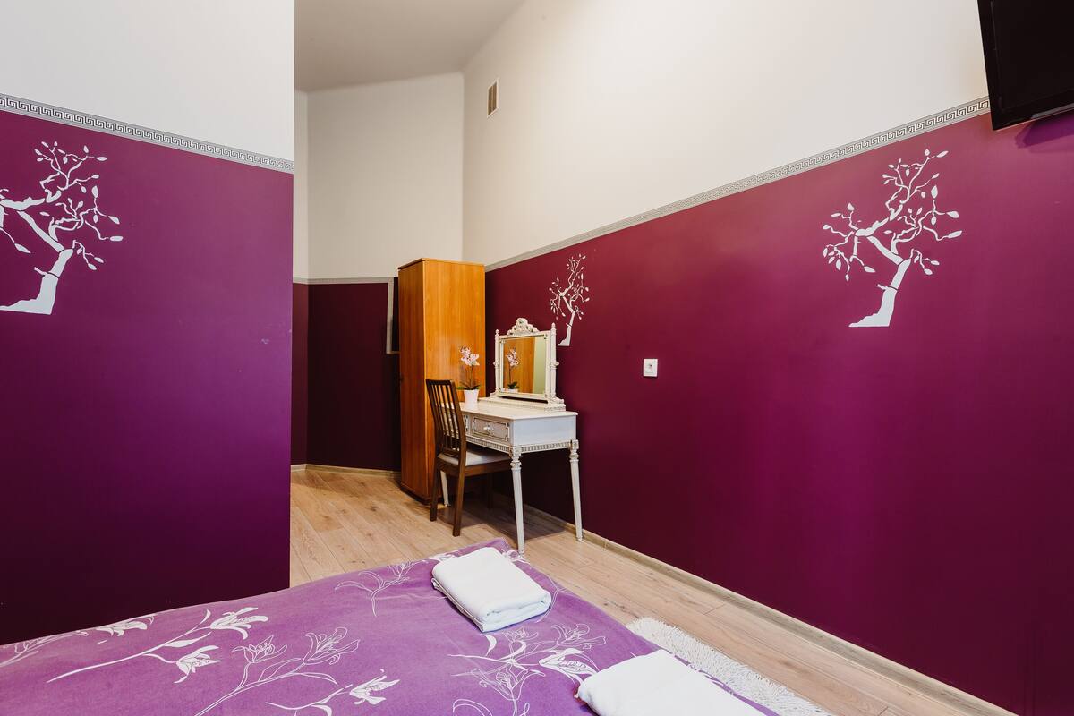 Ein Schlafzimmer mit violetten Wänden, einem Bett mit floralem Überwurf, einem hölzernen Schminktisch und einem Spiegel.