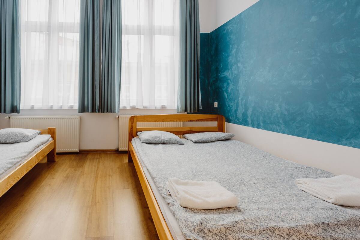 Ein Zimmer mit zwei Einzelbetten, einem Heizkörper und einem großen Fenster mit durchsichtigen Vorhängen.