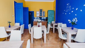Un espace repas avec des tables en bois et des chaises blanches, une cuisine à l’arrière-plan et un mur bleu agrémenté d’éléments décoratifs blancs.