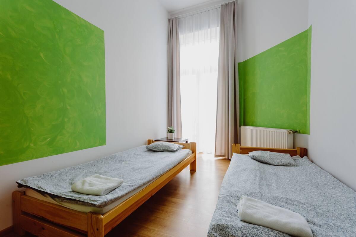 Ein Zimmer mit zwei Einzelbetten, einem kleinen Tisch, einem Stuhl und einem Fenster mit Vorhängen.