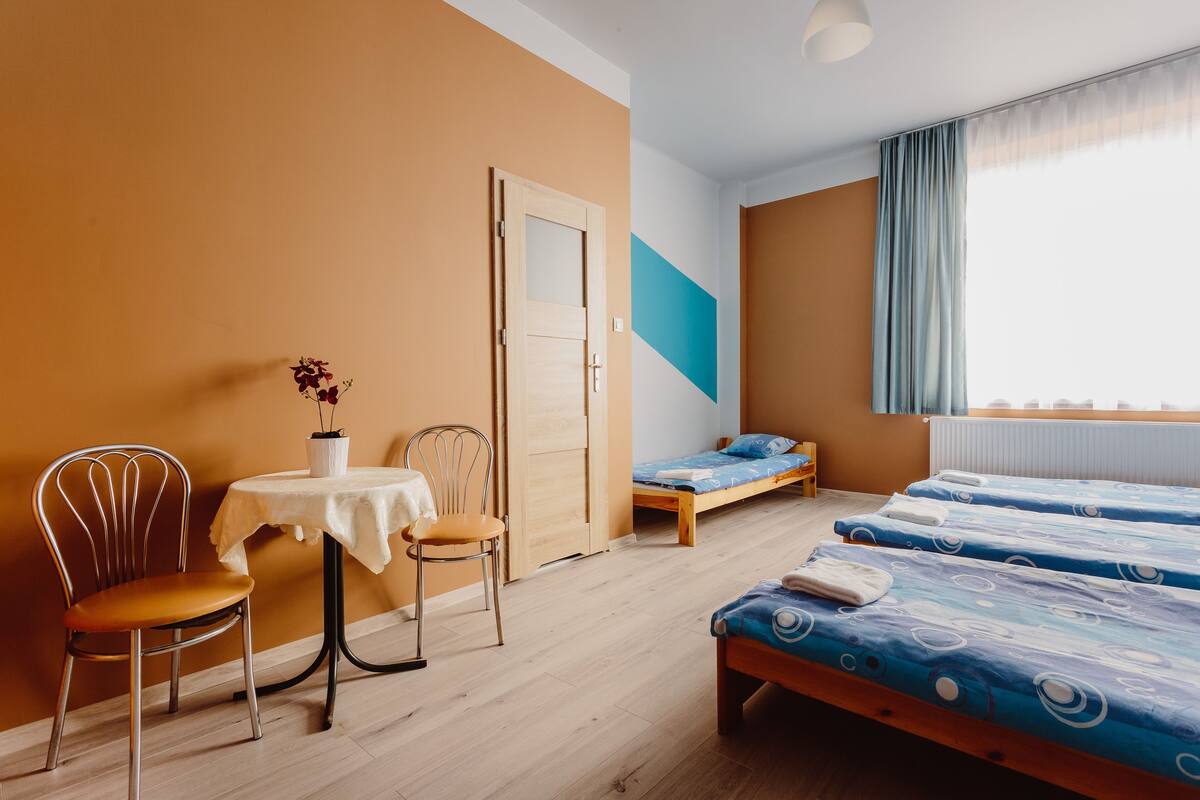 Ein Zimmer mit einem Bett, einem Sessel, einem kleinen Tisch mit einer Vase und einem Fenster mit Vorhängen.