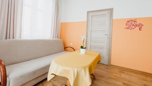 Une pièce avec un canapé beige, une petite table recouverte d’une nappe jaune, un vase blanc contenant une fleur et une porte en bois.