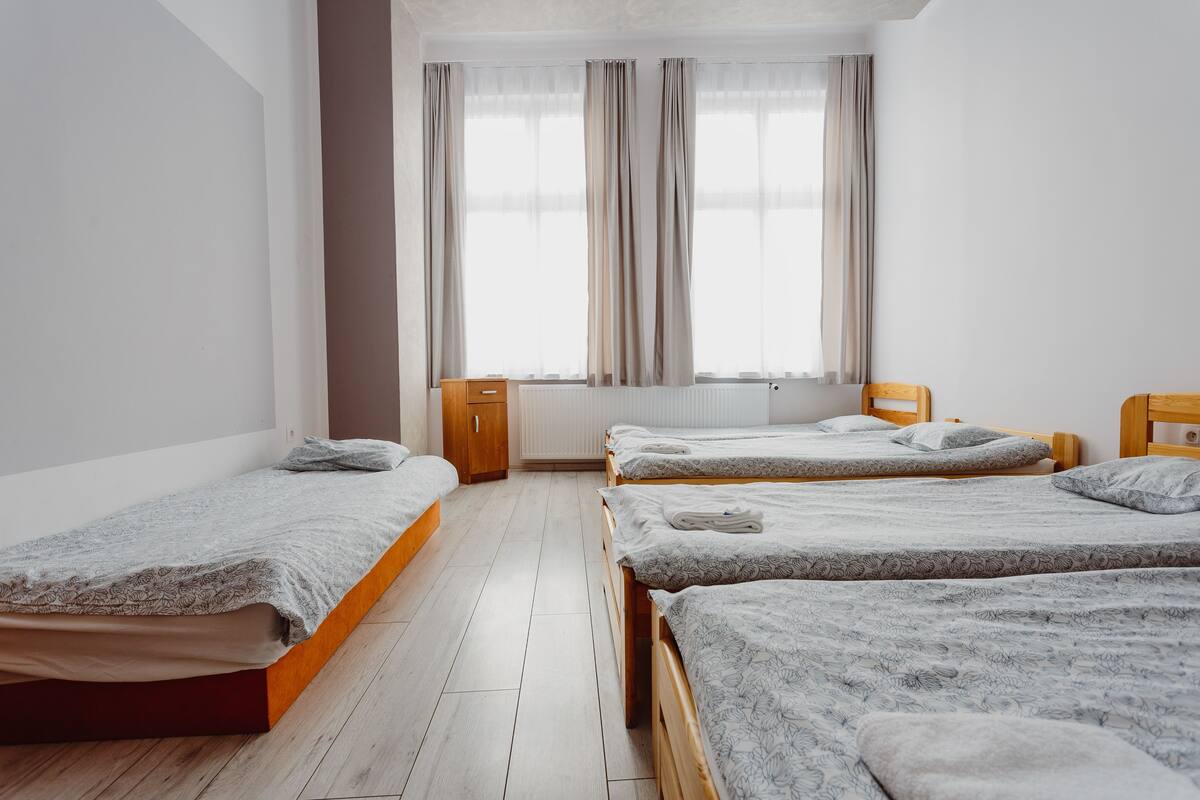 Ein Zimmer mit drei an einer Wand angeordneten Holzbetten, einem Heizkörper und einem fenster mit Vorhängen.