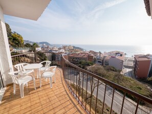 Un balcón con sillas de plástico blanco y piso de baldosas, con vista a un paisaje urbano costero con edificios y el mar.