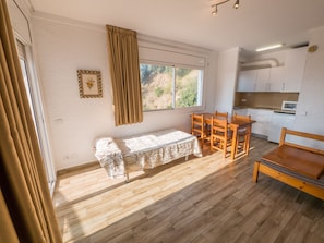 Une pièce avec un plancher en bois, une table à manger avec des chaises, un banc, une fenêtre avec des rideaux, un coin cuisine et un tableau encadré au mur.