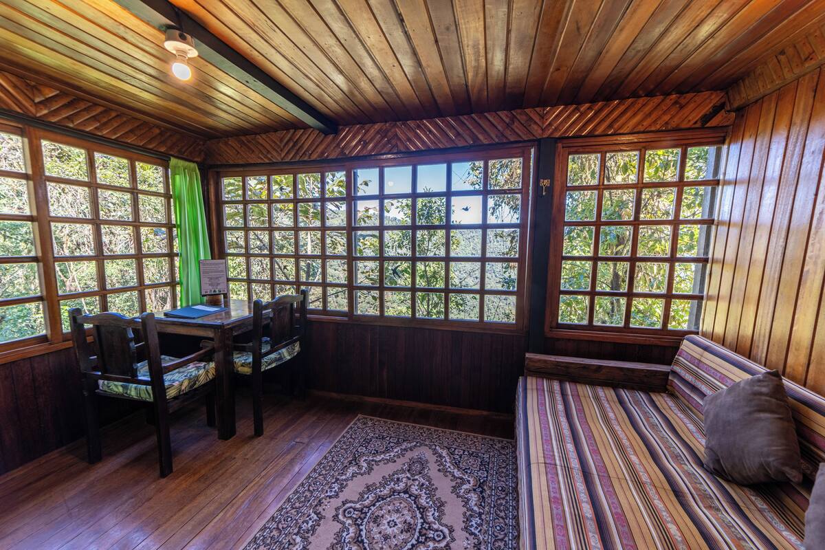 Una habitación con paredes y techo de madera, ventanas amplias con vistas a árboles, una mesa de madera con sillas, y un banco a rayas.