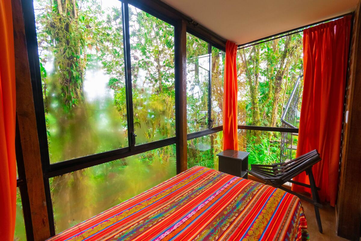 Una habitación con un ventanal grande, una cama con colcha a rayas de colores, una mesita y una silla.