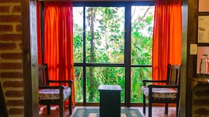 Una habitación con vistas a una vegetación exuberante a través de amplios ventanales con cortinas rojas, dos sillas de madera y una pequeña mesa.