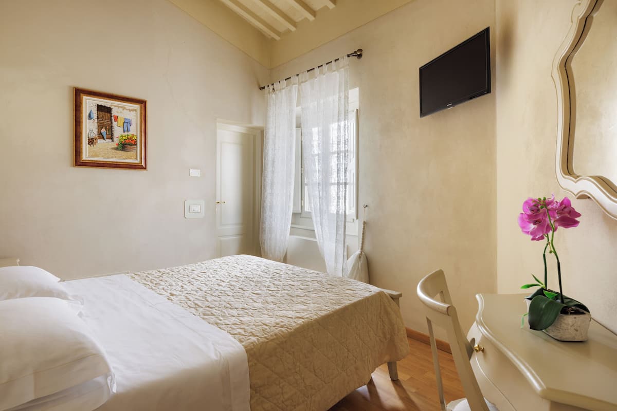 A bedroom with a bed, a chair, a desk, a TV, a window with curtains, and a framed picture on the wall.