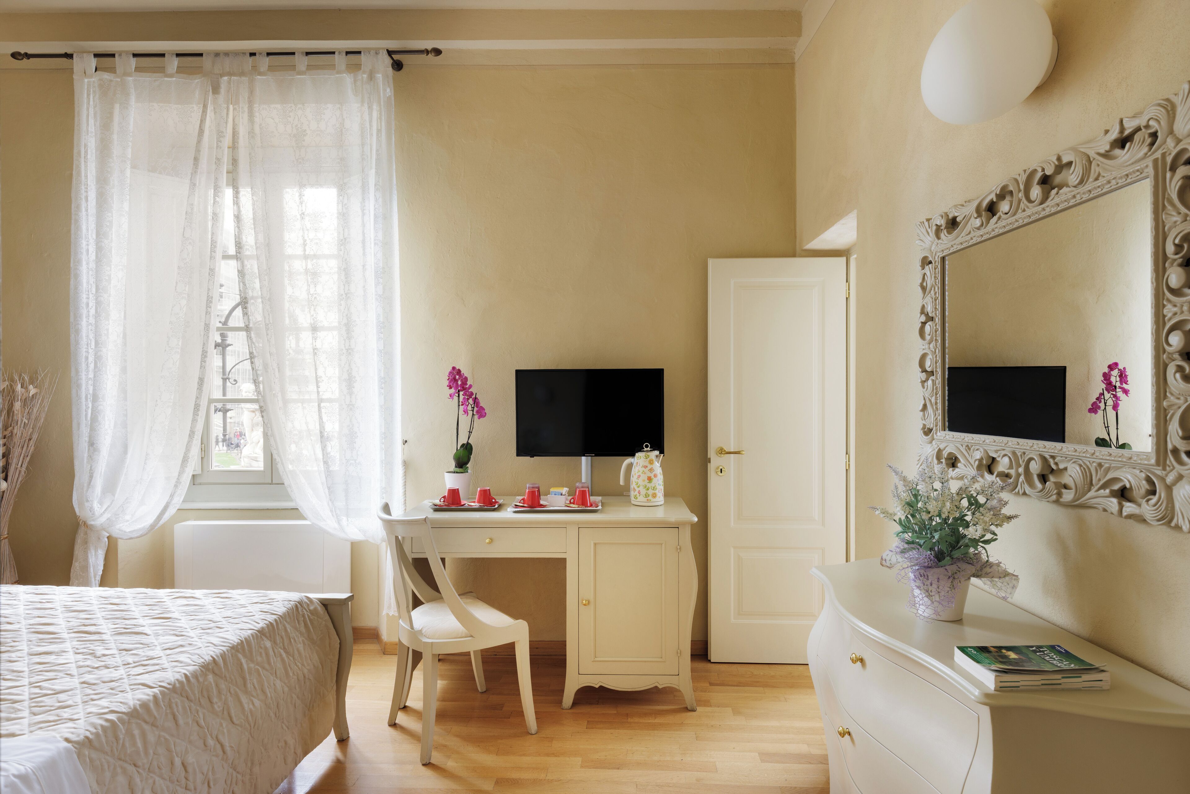 Une chambre à coucher comprenant un lit, un bureau, une chaise, une commode, un miroir et une fenêtre avec des rideaux légers.