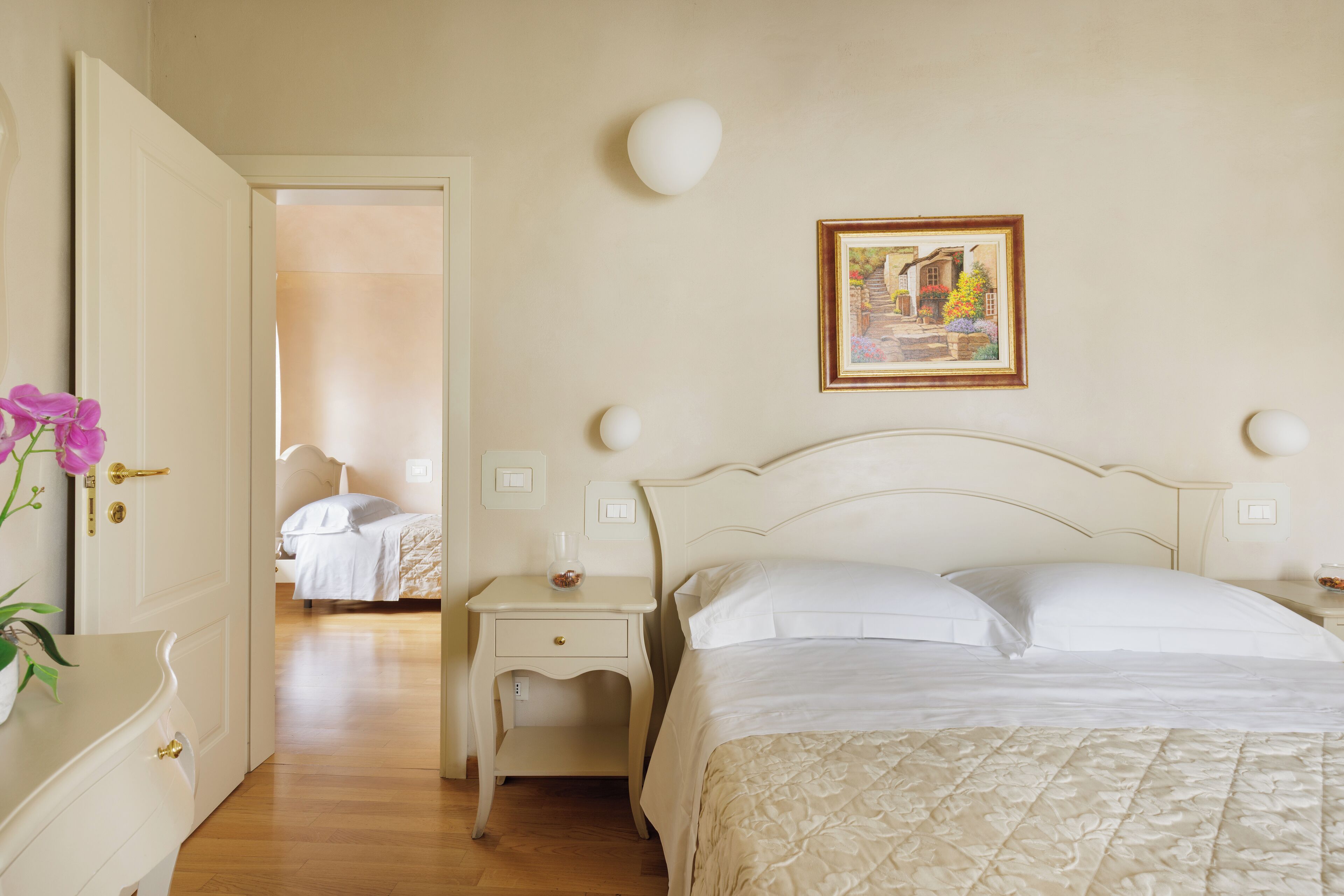 A bedroom with a bed, bedside tables, a nightstand, a painting on the wall, and a door leading to another room.